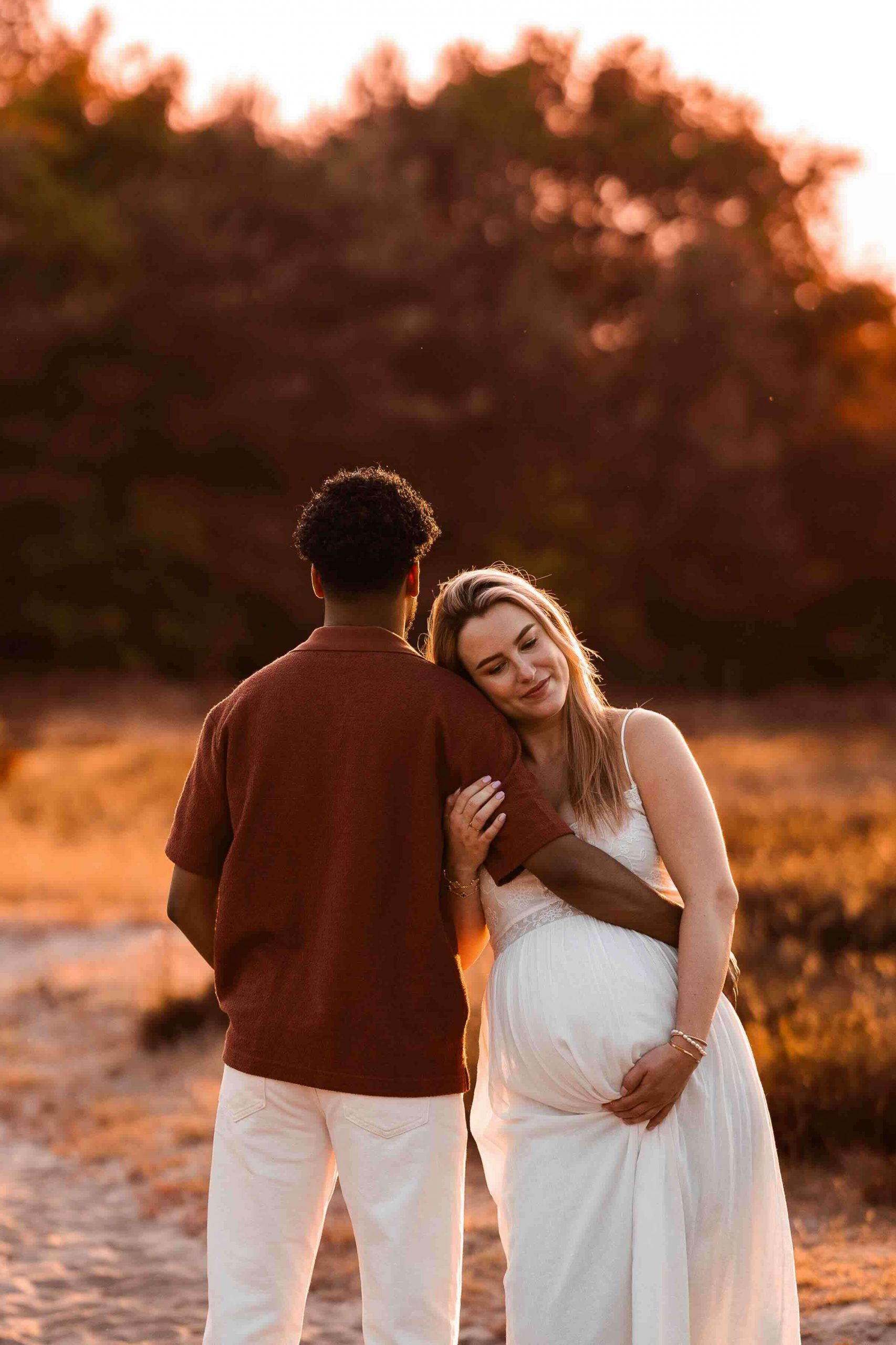 Bruid in witte jurk leunt liefdevol tegen haar partner tijdens een fotoshoot bij zonsondergang in de natuur